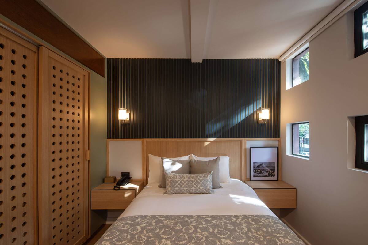 Una habitación de hotel moderna con una cama grande, cabecera de madera, dos mesitas de noche y un gran ventanal.