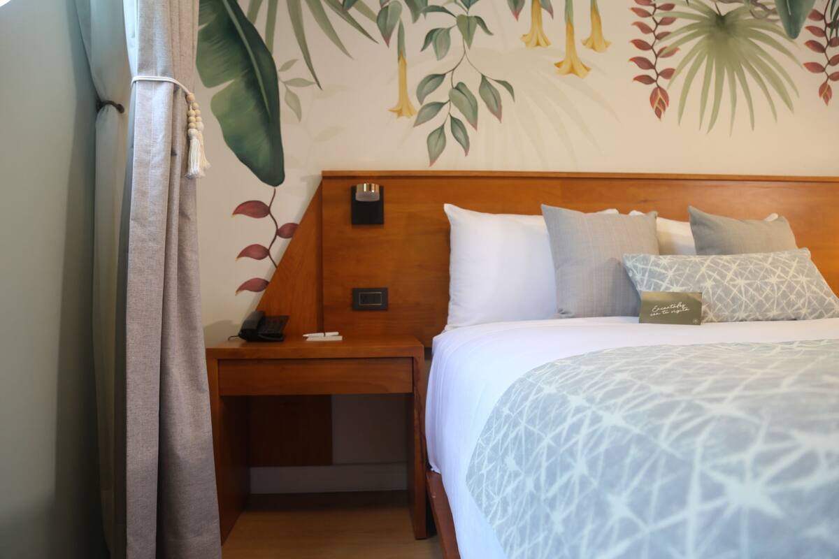 Una cama bien hecha con una colcha estampada, una mesita de noche de madera con un teléfono y un objeto pequeño encima, y una pared con un mural floral.