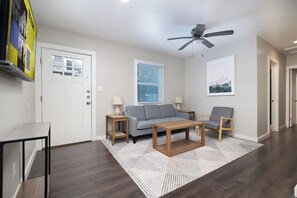 Ein modernes Wohnzimmer mit grauem Sofa, einem Holztisch und einem grauen Sessel. Es gibt einen Deckenventilator, ein Fenster mit Jalousien und eine Tür mit einem Plakat.