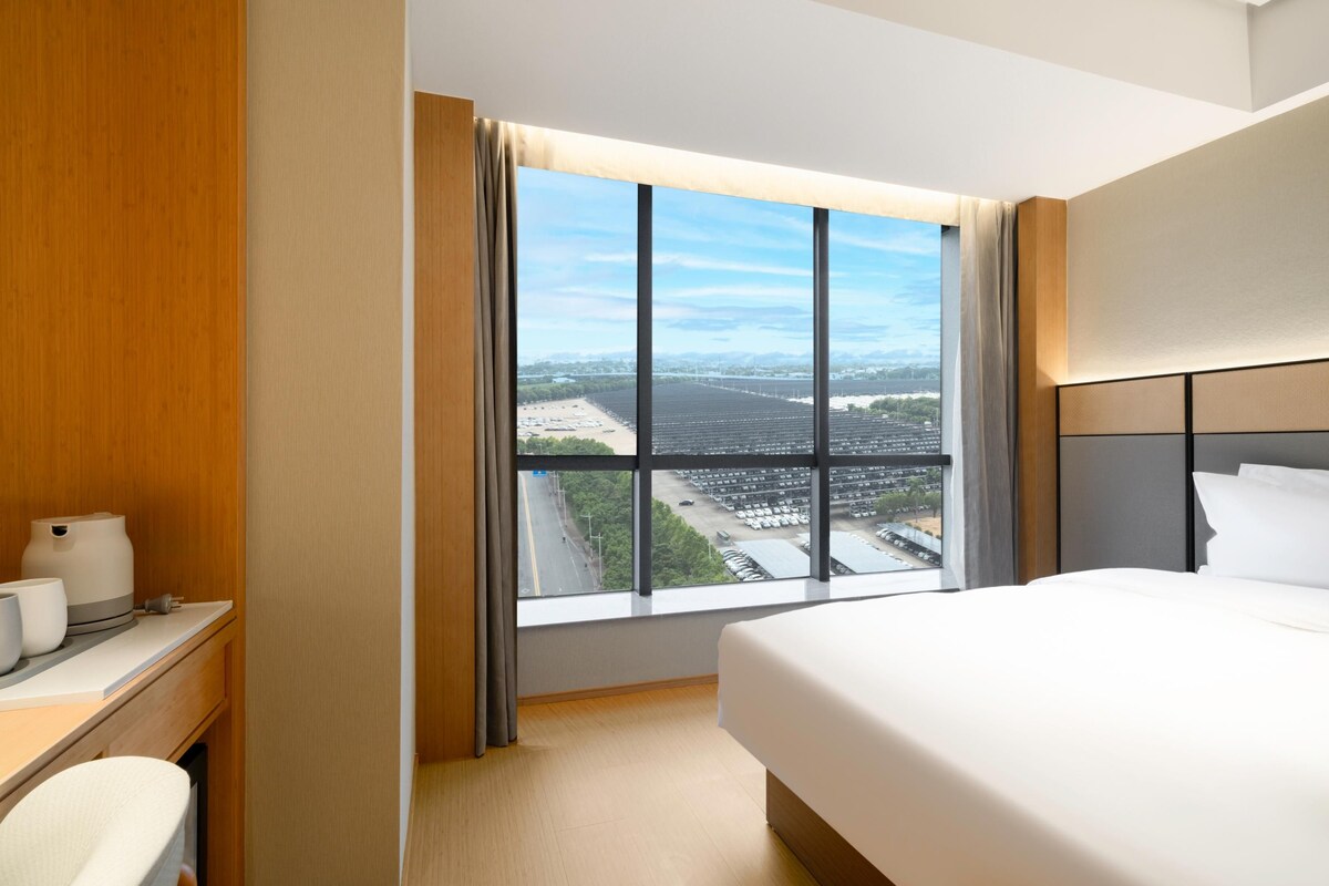 A hotel room with a large window, a bed, a bedside table with a lamp, and a wooden wardrobe.