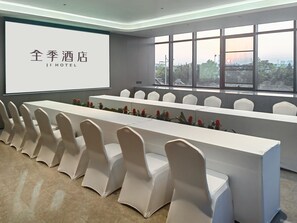 A conference room with a long table and chairs, a projector screen with text, and large windows with a view of buildings and trees. - Ji Hotel Guangzhou North Railway Station (Guangzhou)