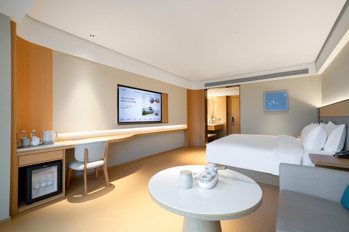 Ein Hotelzimmer mit einem Bett, einem Schreibtisch, einem Sessel, einem kleinen Kühlschrank, einem Tisch mit Teekannen und einem an der Wand befestigten Fernseher.