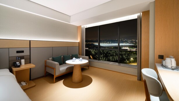 A modern hotel room with a large window offering a nighttime city view, a sofa, a small round table, and a desk with a chair.