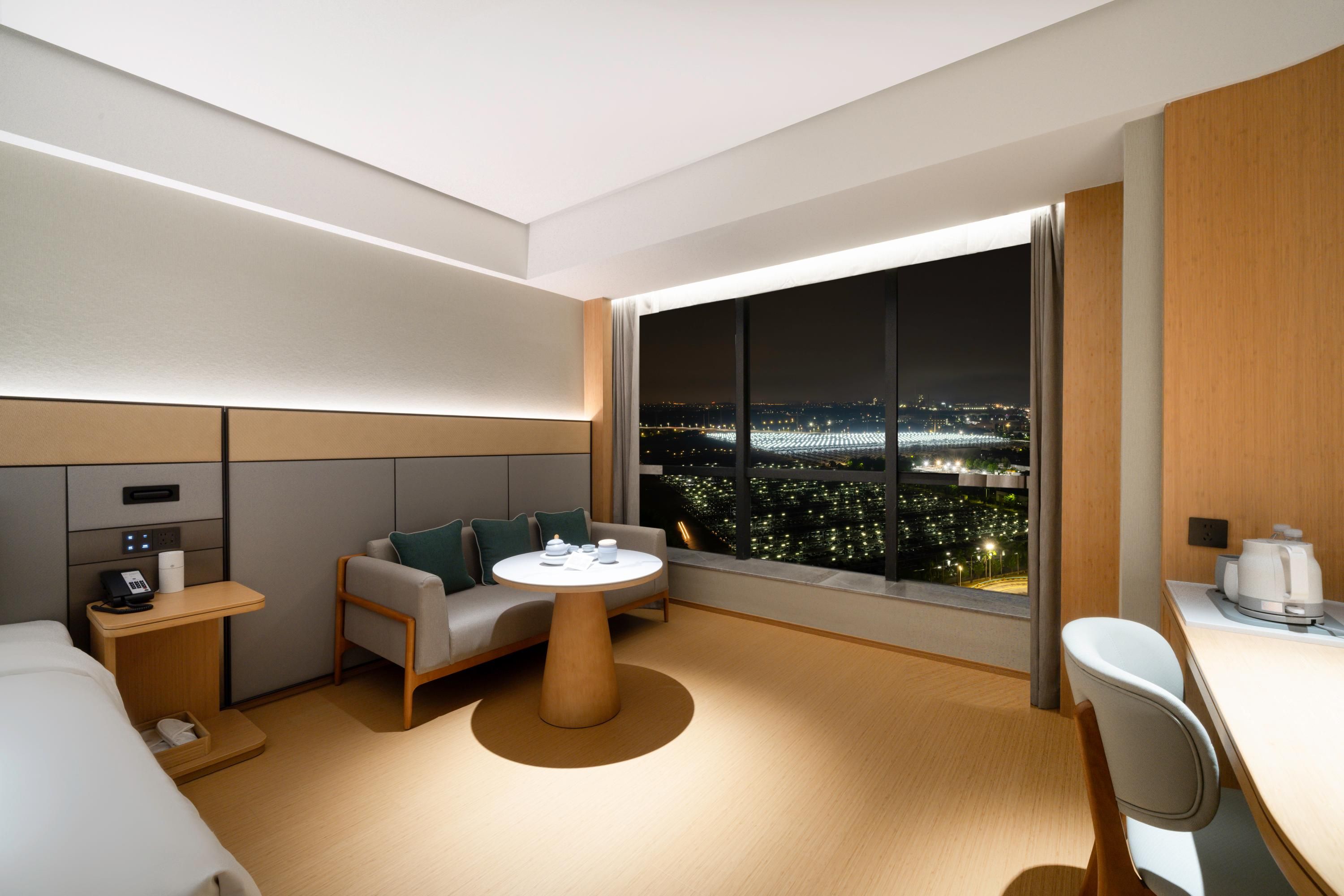 Habitación de hotel moderna con un amplio ventanal que ofrece vistas nocturnas de la ciudad, un sofá, una pequeña mesa redonda y un escritorio con una silla.