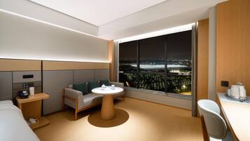 Habitación de hotel moderna con un amplio ventanal que ofrece vistas nocturnas de la ciudad, un sofá, una pequeña mesa redonda y un escritorio con una silla.