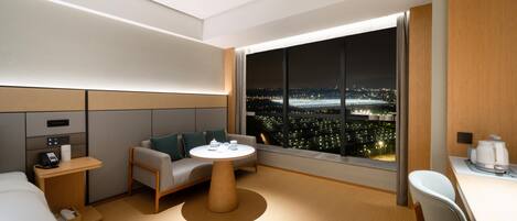 Une chambre d’hôtel moderne dotée d’une grande fenêtre offrant une vue sur la ville la nuit, d’un canapé, d’une petite table ronde et d’un bureau avec une chaise.