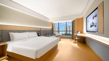 A modern hotel room with a large bed, a desk, and a view of the cityscape through large windows.