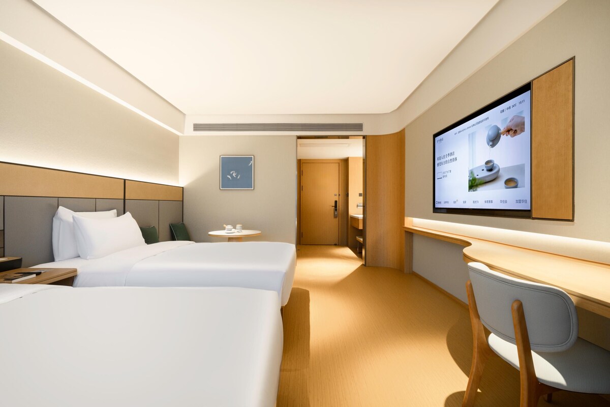 A hotel room with two beds, a desk, a chair, a TV mounted on the wall, and a small table with a cup.