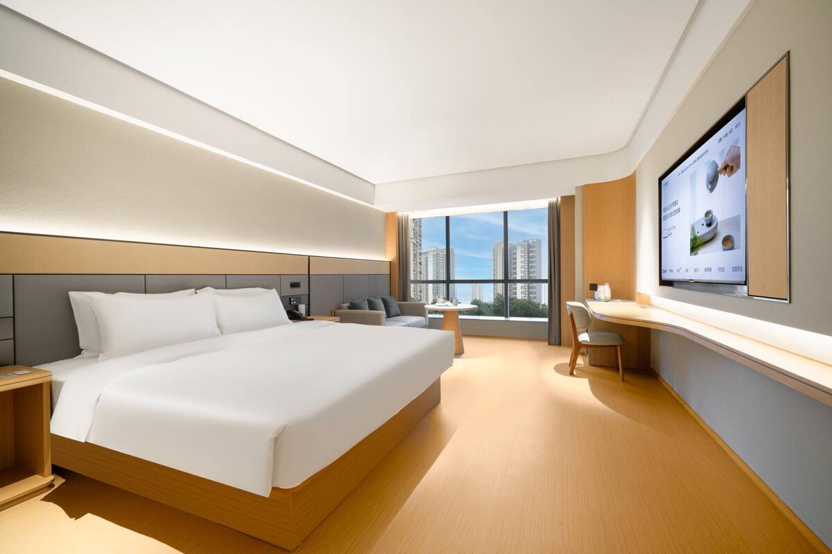 Ein modernes Hotelzimmer mit einem großen Bett, einem Schreibtisch und Blick auf die Stadt durch große Fenster.