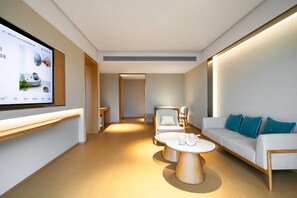 A modern hotel room with a flat-screen TV mounted on the wall, a sofa with blue cushions, a round coffee table, and a seating area with chairs. - Ji Hotel Guangzhou North Railway Station (Guangzhou)