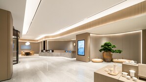 A modern hotel lobby with a reception desk, digital information screen, seating area, and a large potted plant.