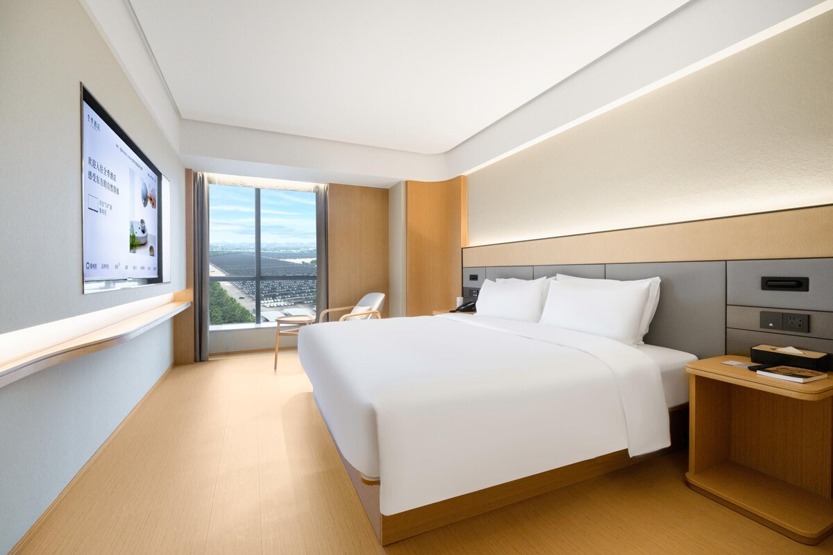 Ein modernes Hotelzimmer mit einem großen Bett, einem Nachttisch, einem Sessel, einem an der Wand befestigten Fernseher und einem Fenster mit Blick auf die Stadt.