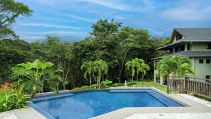 Una piscina rectangular con borde de azulejos, rodeada de abundante vegetación y una casa con balcón.