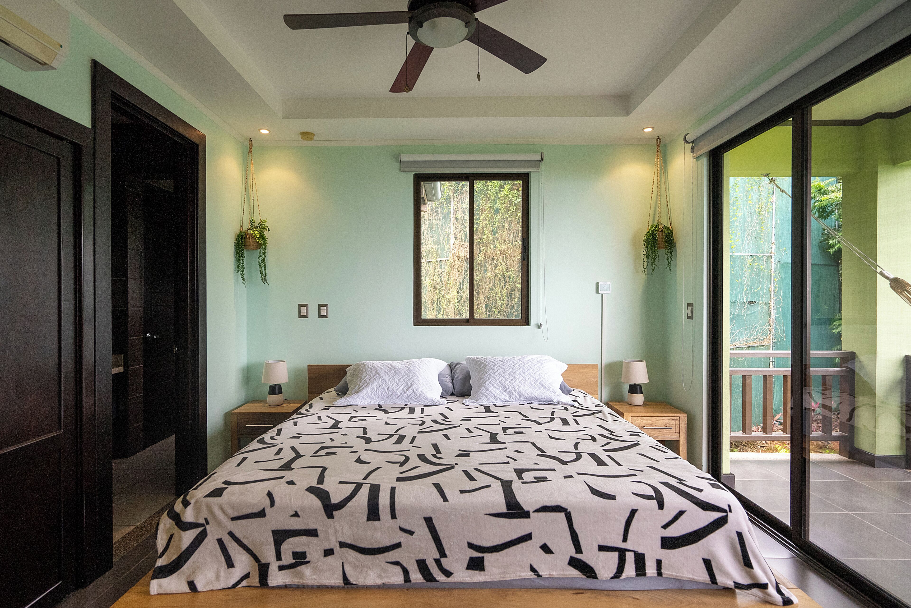 A bedroom with a bed, two bedside tables, a ceiling fan, a window with a view, and a sliding glass door to a balcony.