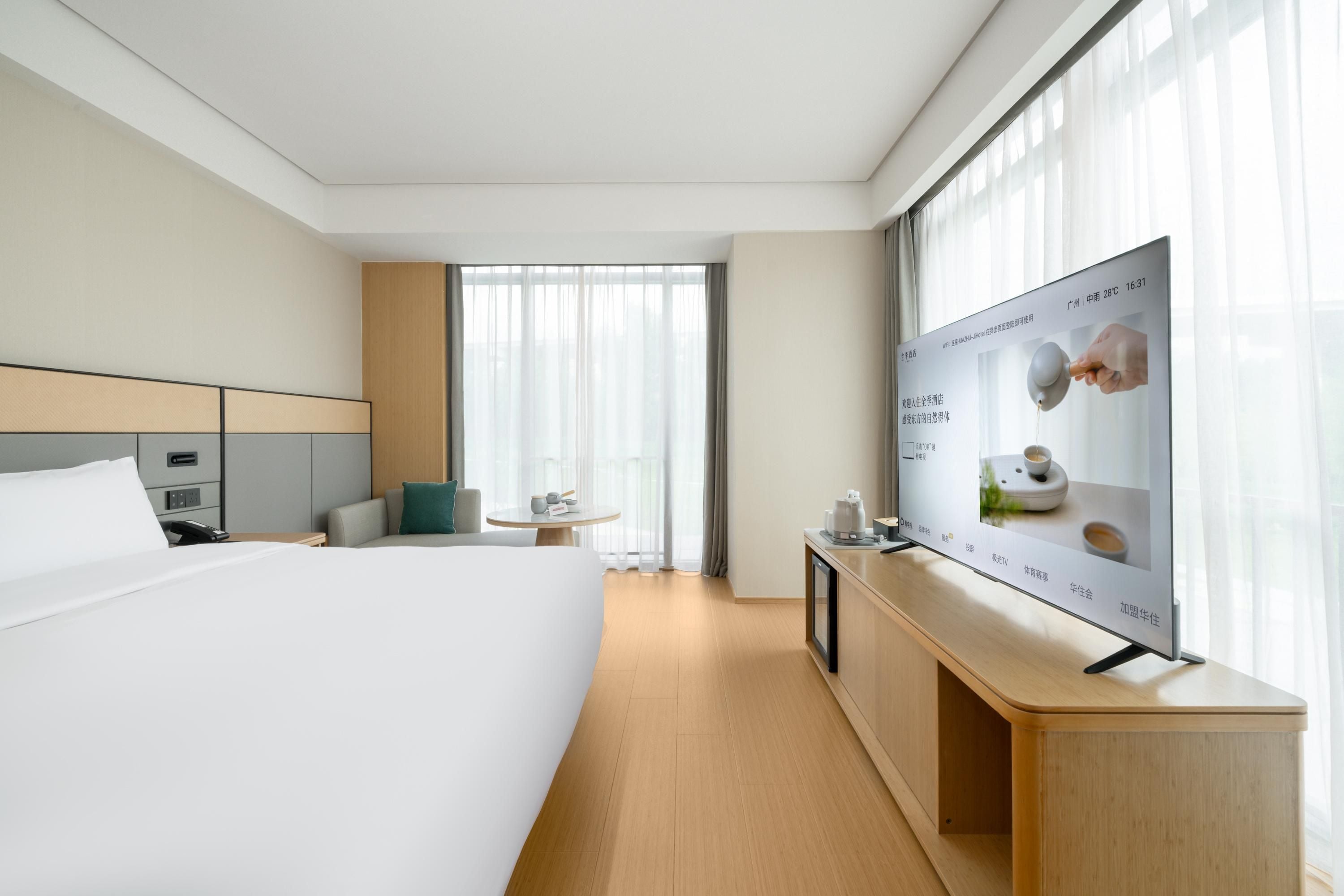 A hotel room with a large bed, a bedside table with a lamp, a small round table with a chair, and a large window with curtains.