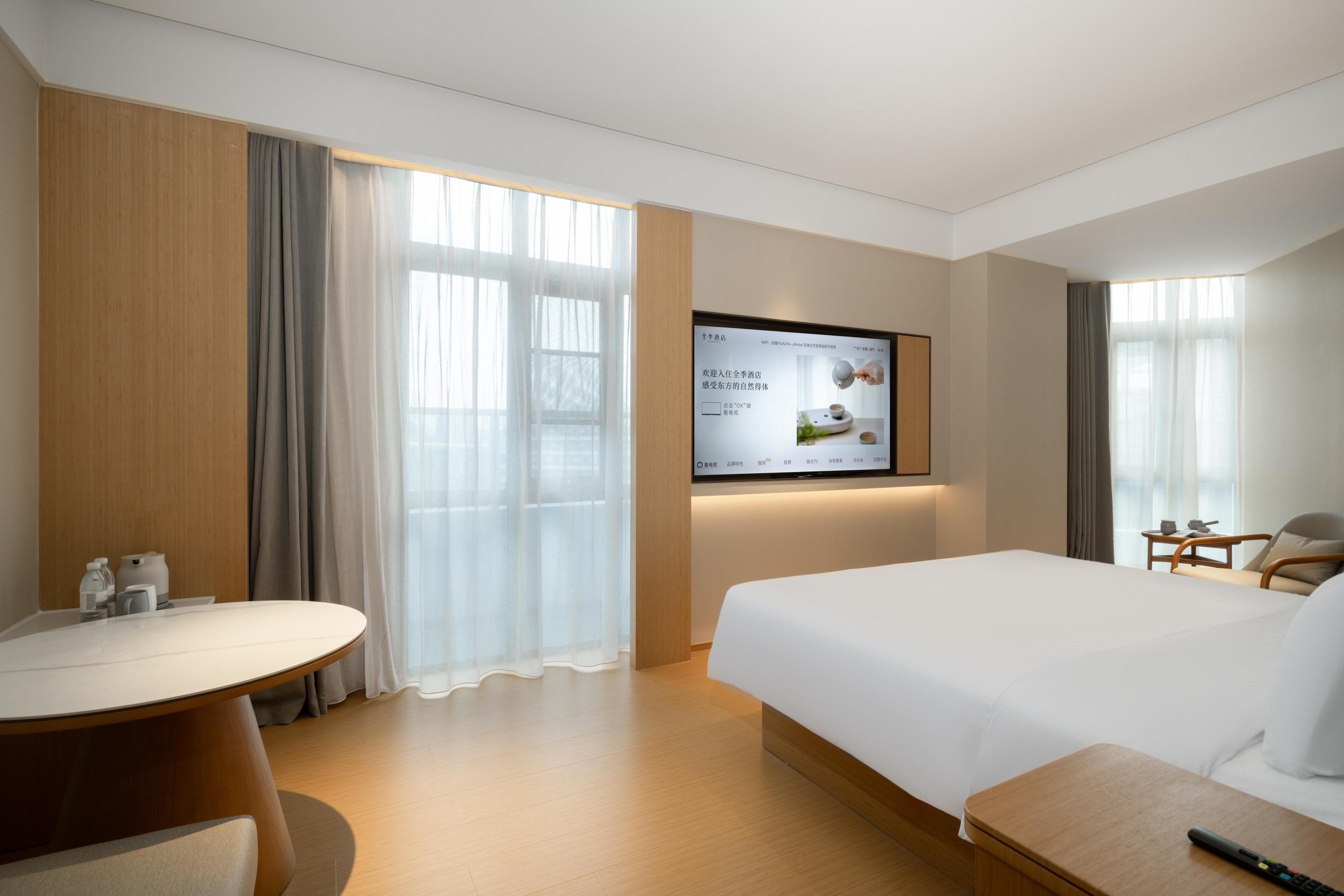 A hotel room with a bed, a desk, a TV mounted on the wall, and a window with curtains.