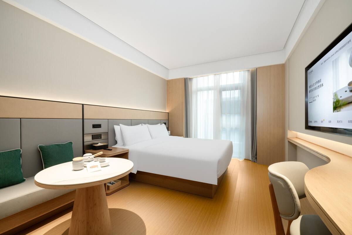 A modern hotel room with a large bed, a bedside table, a small desk, a chair, and a TV mounted on the wall.