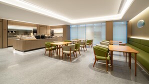 A modern dining area with green upholstered booths and wooden tables, a kitchen in the background, and a large window with a view. - Ji Hotel Guangzhou Tianhe Smart City Wetland Park (Guangzhou)