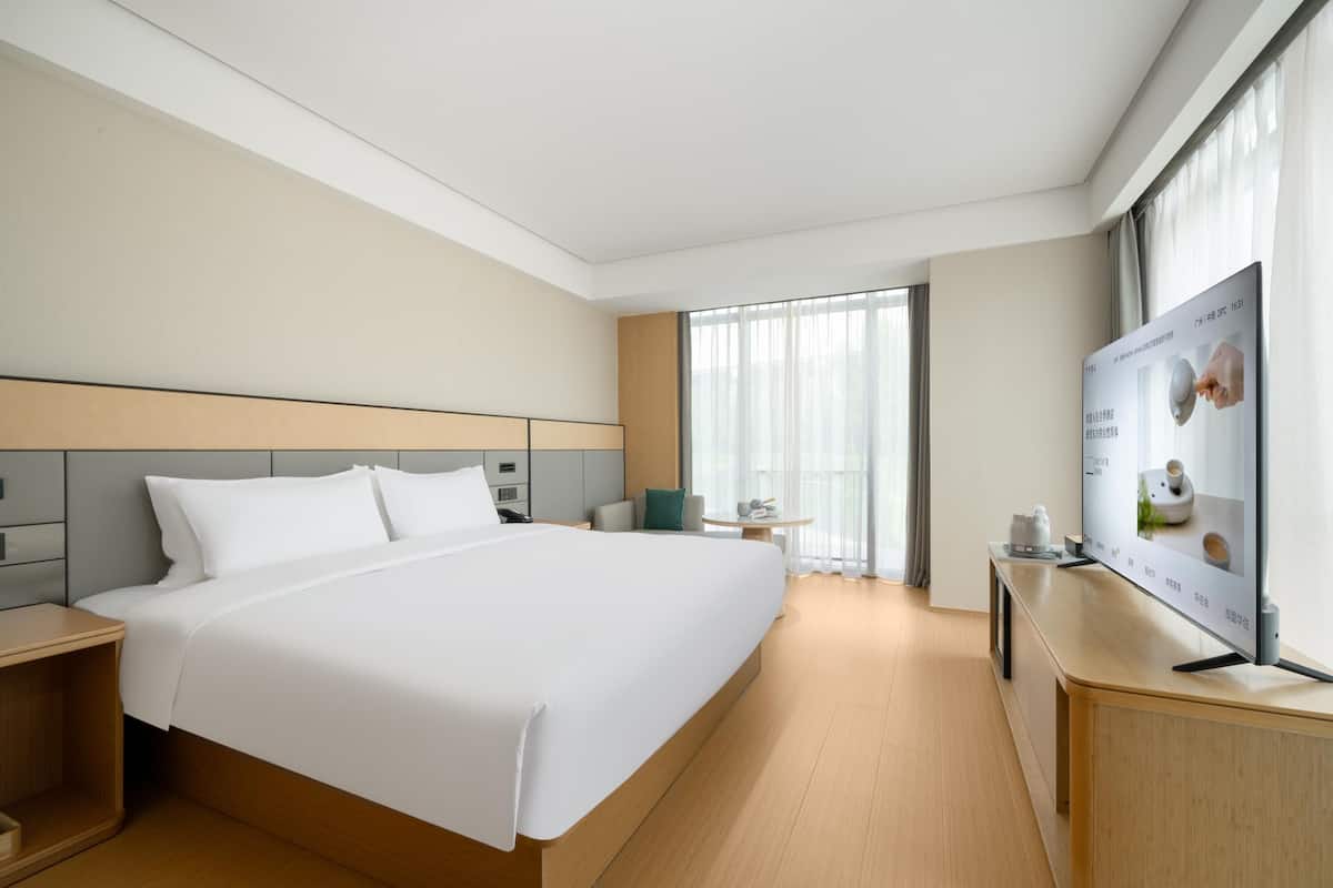 A modern hotel room with a large bed, a bedside table, a desk with a flat-screen TV, and a window with curtains.