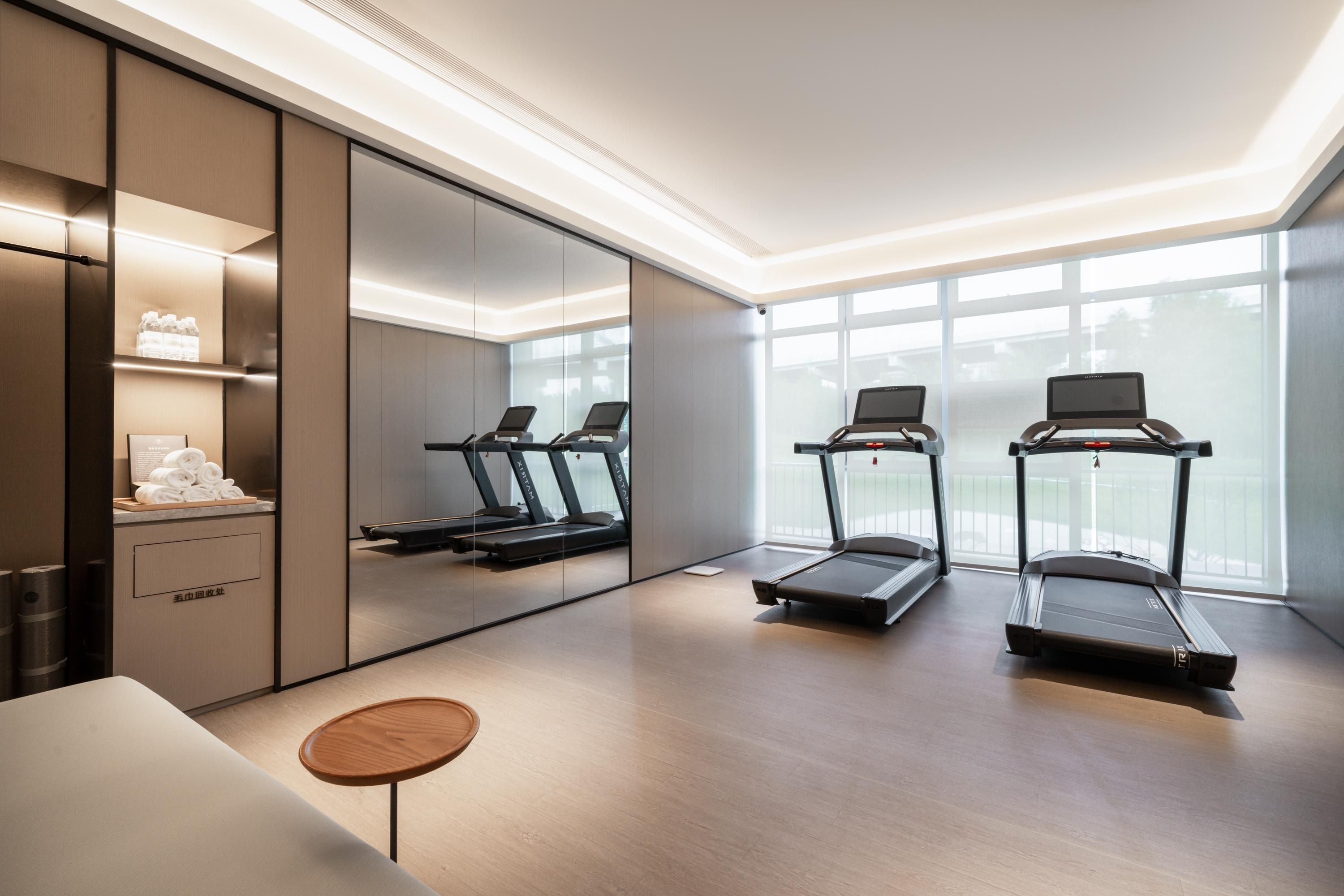 Una habitación de hotel moderna con un espejo grande, dos cintas de correr, una pequeña mesa de madera y vistas de la ciudad a través de amplias ventanas.