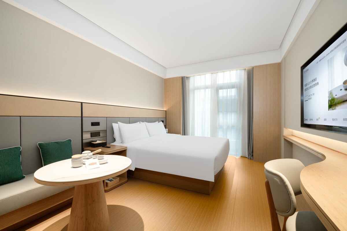 Ein modernes Hotelzimmer mit einem großen Bett, einem Nachttisch, einem kleinen Schreibtisch, einem Stuhl und einem an der Wand befestigten Fernseher.