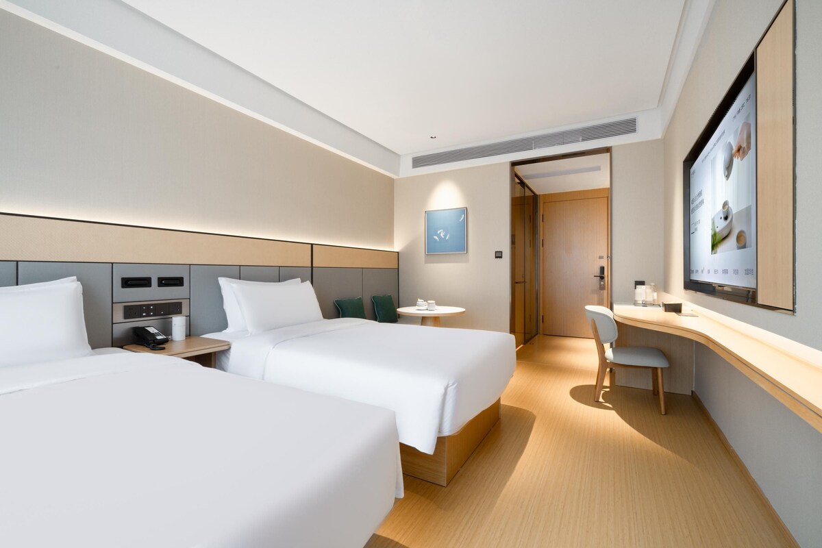 A hotel room with two beds, a desk, a chair, a small table, and a television.