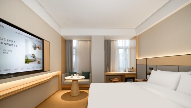A modern hotel room with a large flat-screen TV mounted on the wall, a bed with white linens, a wooden nightstand, a small round table with a chair, and a sofa with a green cushion. - Ji Hotel Guangzhou Tianhe Smart City Wetland Park (Guangzhou)