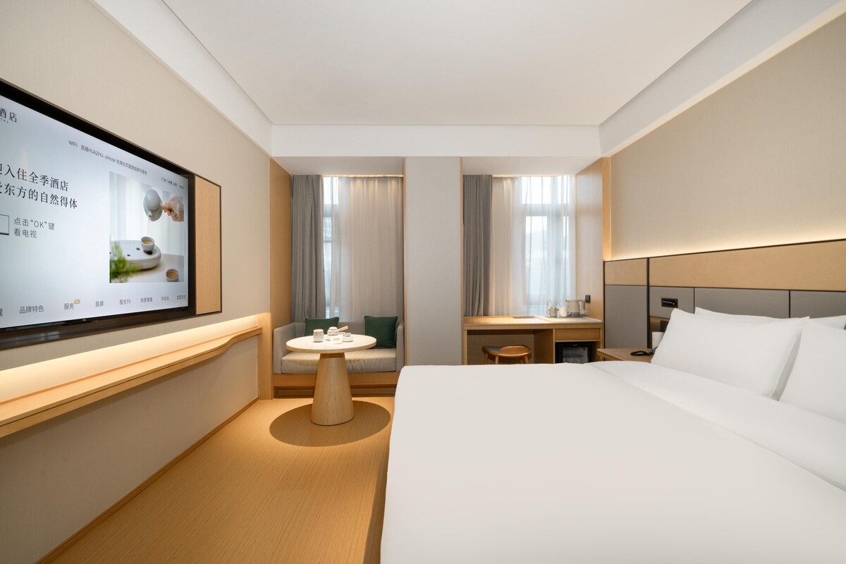 A modern hotel room with a large flat-screen TV mounted on the wall, a bed with white linens, a wooden nightstand, a small round table with a chair, and a sofa with a green cushion.