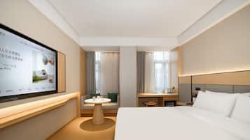 Ett modernt hotellrum med en stor, platt-TV som är monterad på väggen, en säng med vita sängkläder, ett träbord som sängbord, ett litet runt bord med en stol och en soffa med en grön kudde.