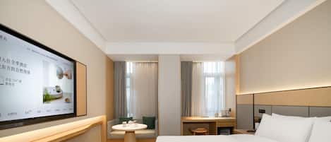 A modern hotel room with a large flat-screen TV mounted on the wall, a bed with white linens, a wooden nightstand, a small round table with a chair, and a sofa with a green cushion.