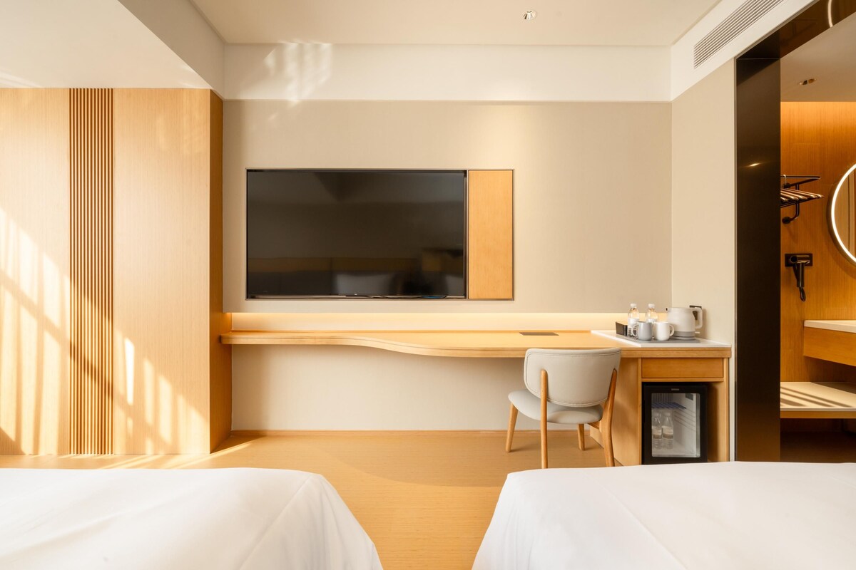A hotel room with a large flat-screen TV mounted on the wall, a small desk with a chair, a mini-fridge, and a bed with a wooden headboard.