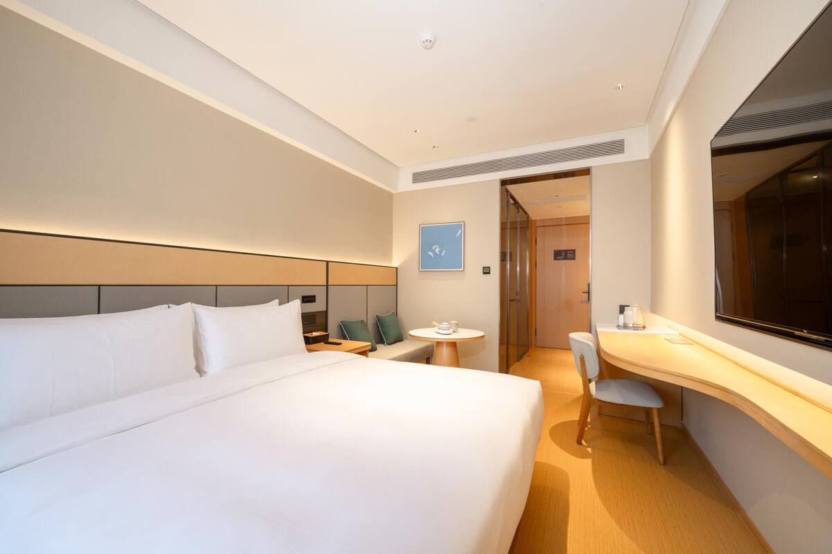 A modern hotel room with a large bed, a desk, and a chair. There is a small table with a cup and a book. The room has a large window and a door leading to another room.