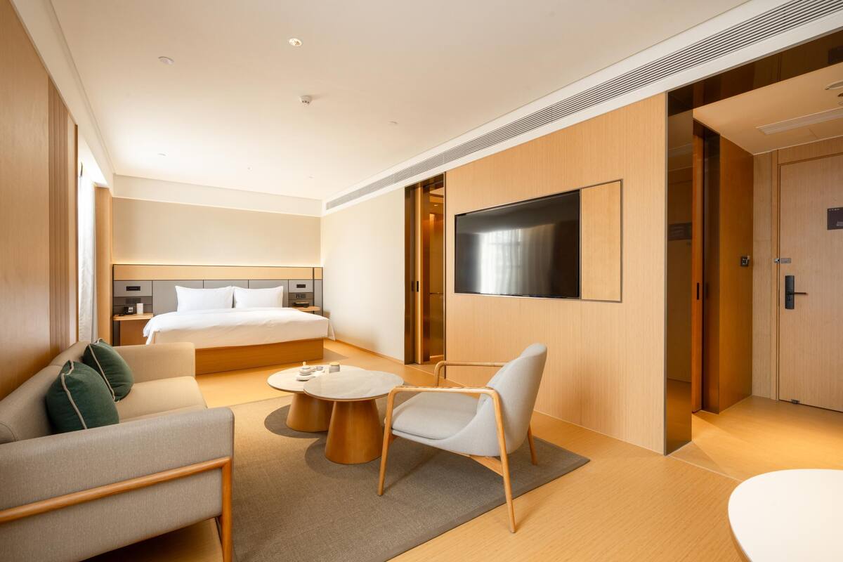 A modern hotel room with a large bed, a sofa, a chair, a TV, and a small table.
