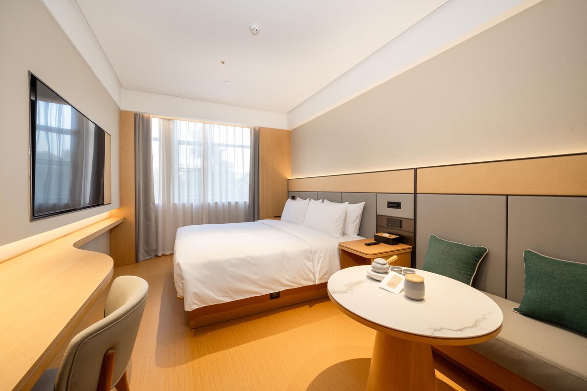 A modern hotel room with a large bed, bedside tables, a desk, and a round table with a vase.
