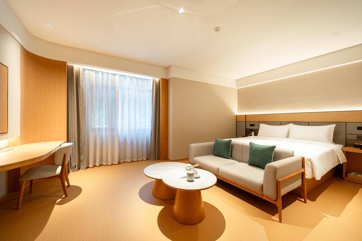 A modern hotel room with a bed, a sofa, a desk, and two round tables.