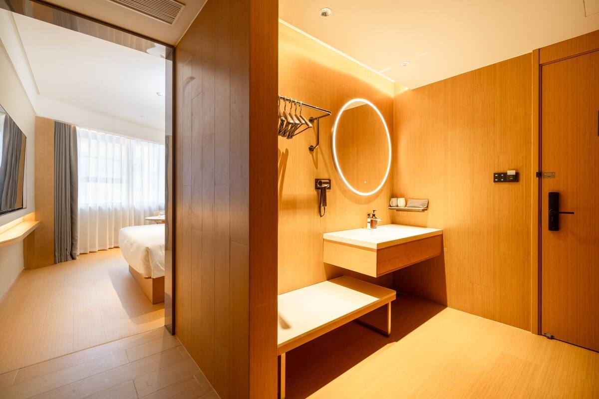 A modern hotel room with a wooden floor, a bed, a bathroom with a sink and mirror, and a wardrobe.
