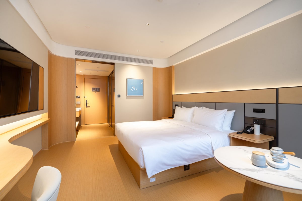 A modern hotel room with a large bed, bedside table, and a small round table with a lamp.