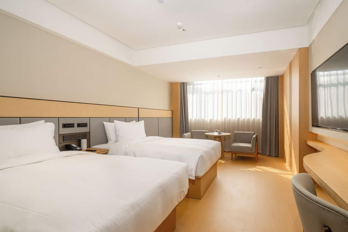 A hotel room with two beds, a small table, a chair, a TV, and a window with curtains.