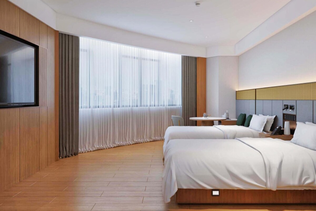 A hotel room with two beds, a TV, a small table, and a window with curtains.