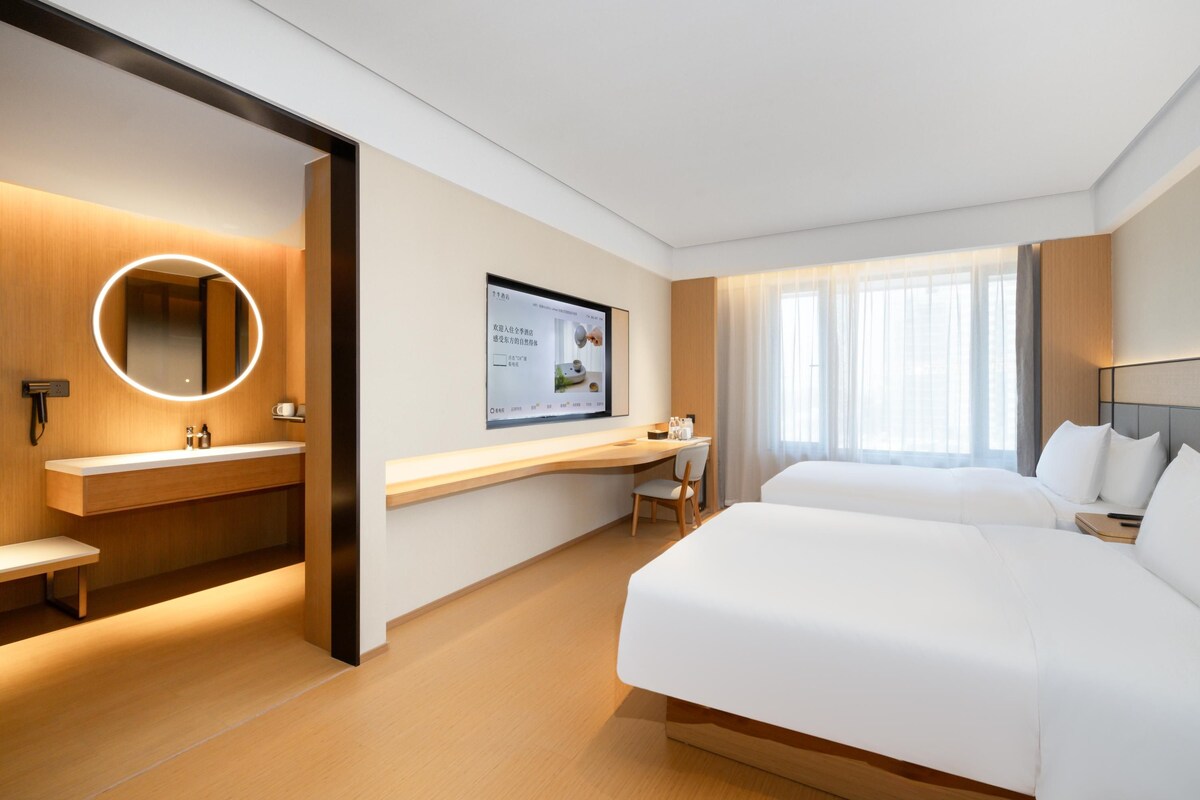 A modern hotel room with a large bed, a desk, a chair, a round mirror, and a TV screen.