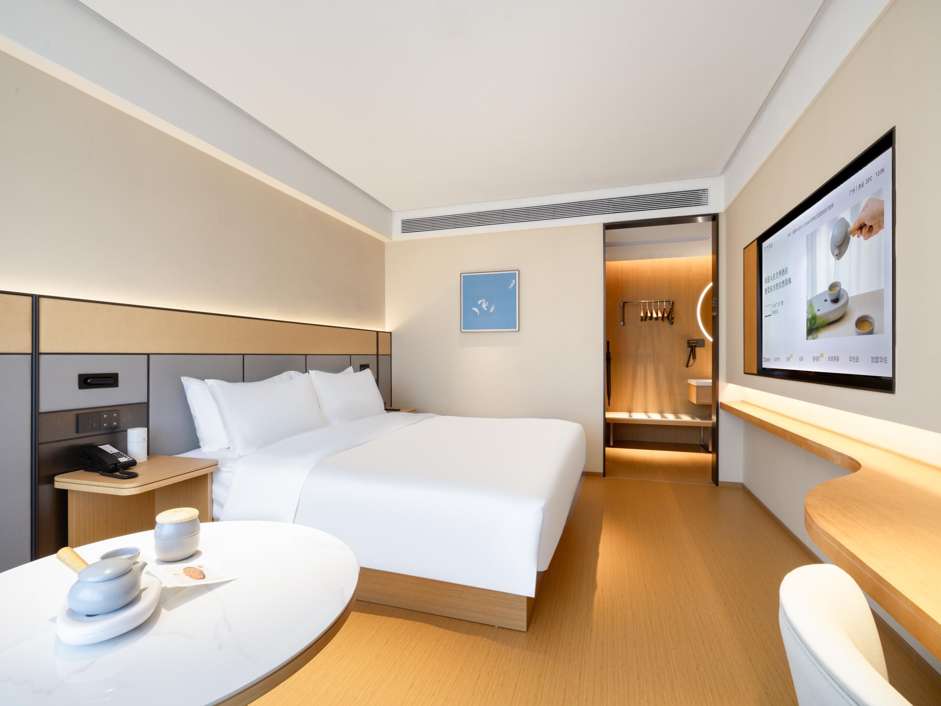 A modern hotel room with a neatly made bed, a bedside table with a phone and a cup, and a wall-mounted TV displaying a menu.