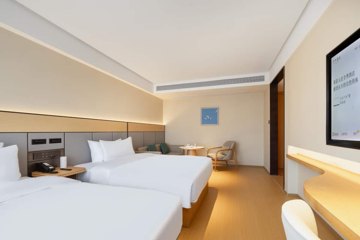 Une chambre d’hôtel avec deux lits, un bureau, une chaise, une petite table et un téléviseur fixé au mur.