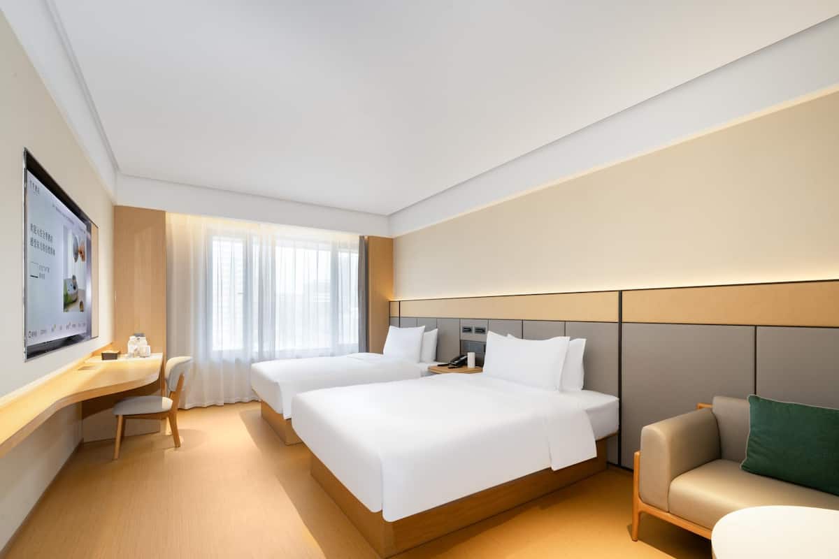 Une chambre d’hôtel avec deux lits, un bureau, une chaise, un canapé et une grande fenêtre avec des rideaux légers.