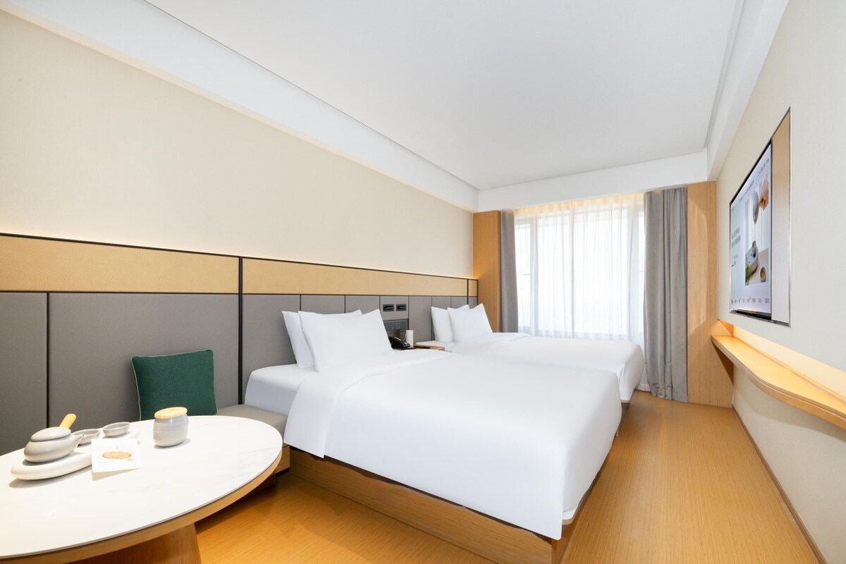 A modern hotel room with a large bed, a bedside table with a lamp, a small seating area, and a large window with curtains.