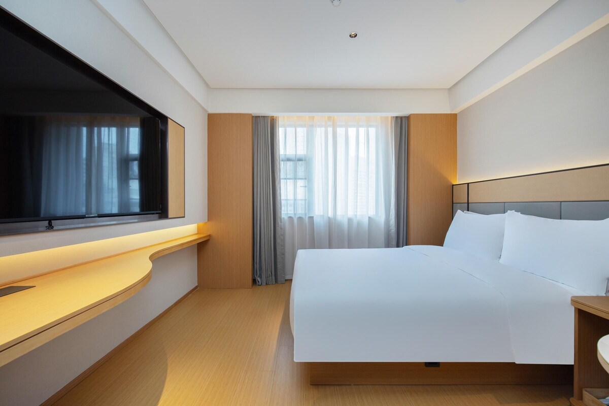 A hotel room with a bed, a TV, a bedside table, and a window with curtains.