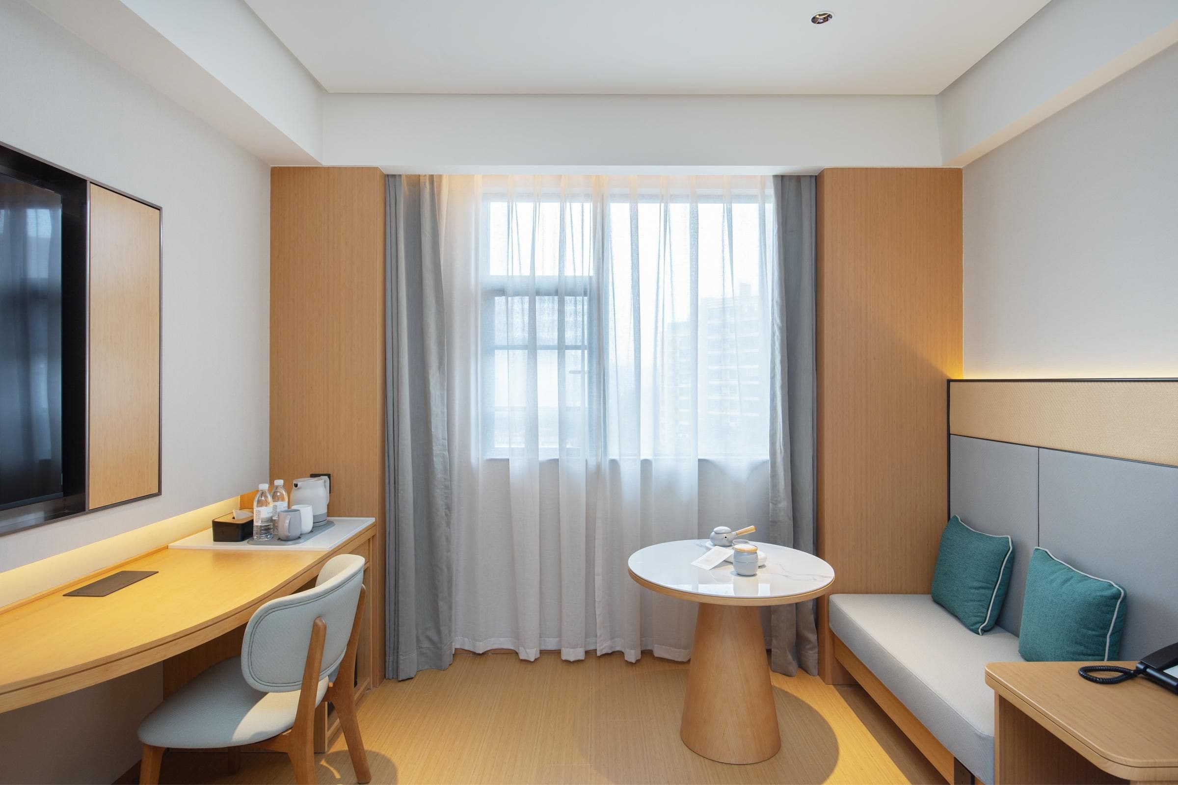 A modern hotel room with a wooden desk, a round table, a sofa, and a large window with sheer curtains.
