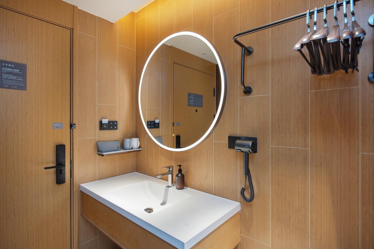 A modern bathroom with a white sink, a round mirror with a light, a towel rack, and a shower area.