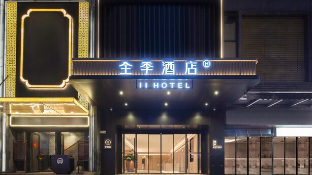 Entrada de um hotel com uma placa indicando "JI HOTEL" e uma grande planta em vaso no interior.
