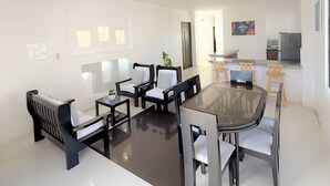 Un salon moderne avec une table à manger au plateau en verre, des chaises noires et blanches, un canapé et un coin cuisine équipé d’un téléviseur et de tabourets.