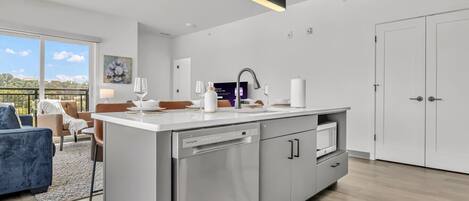 Une cuisine moderne équipée d’un lave-vaisselle, d’un évier et d’un plan de travail. On y trouve également un coin repas avec une table et des chaises, et de grandes fenêtres offrent une vue sur l’extérieur.
